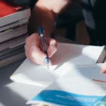 Seseorang sedang menulis catatan di buku tulis dengan tumpukan buku pelajaran di sampingnya